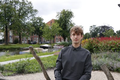 The photo shows a close-up of a young man, Jasper Bitter, in a grey shirt. He is standing in front of a flower bed and smiling slightly at the camera. To the left in the background is a canal with a few boats and behind it some trees and buildings can be seen.