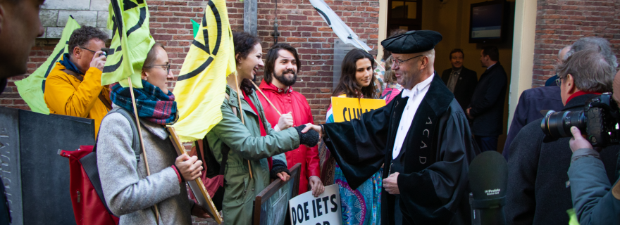The photo shows a group of Extinction Rebellion activists with banners on the left. One of the activists is shaking hands with a professor from Leiden University in a black robe and hat, on the right. A camera points at the group from the lower right corner of the picture. In the background is a brick building with an open door.